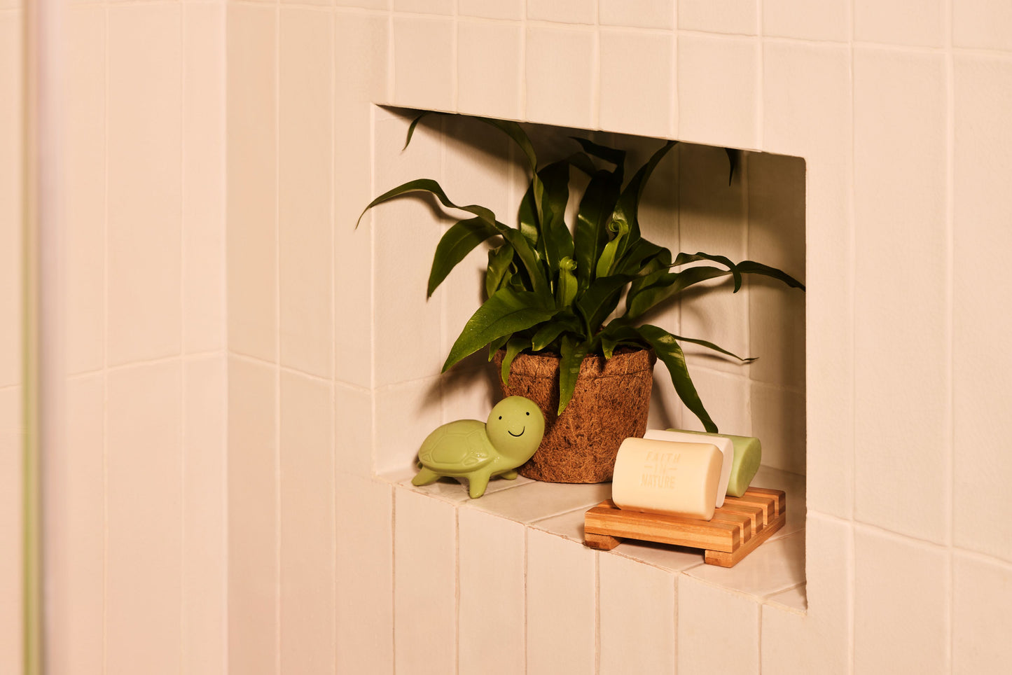 Tre naturlige og veganske sæbebarer placeret på en sæbeholder af træ i en bruseniche ved siden af en grøn plante og en lille dekorativ skildpadde.