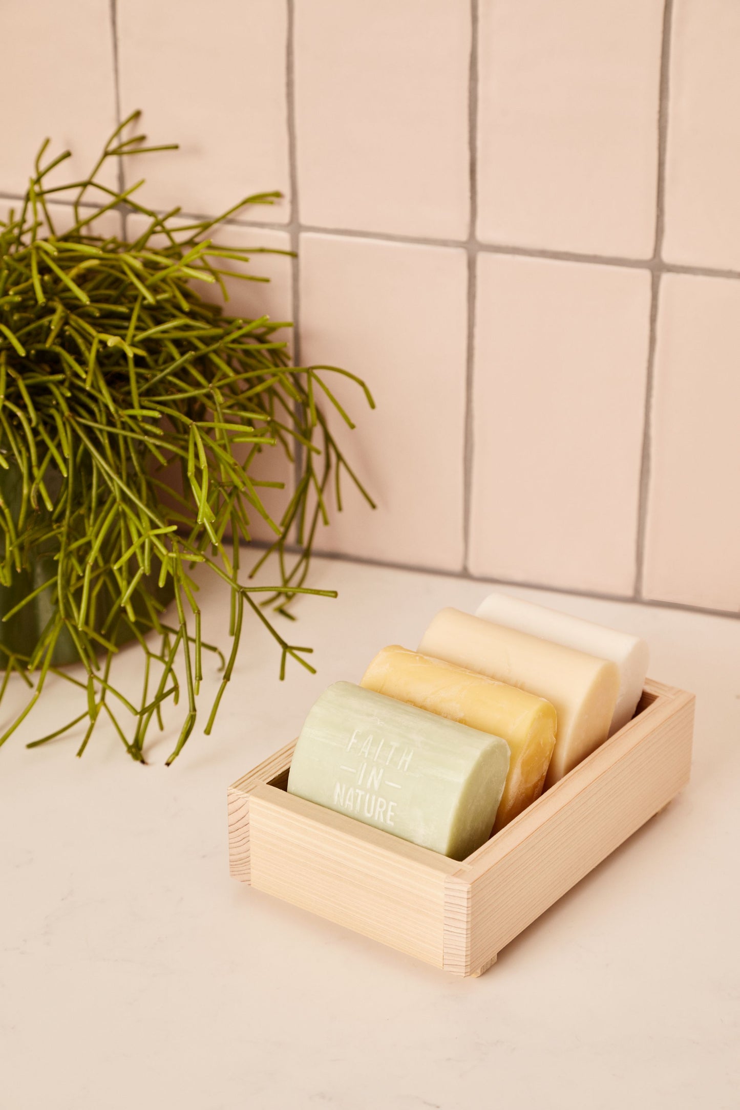 Fire forskellige Faith In Nature sæbebarer uden emballage placeret i en dekorativ træskål ved siden af en grøn plante.