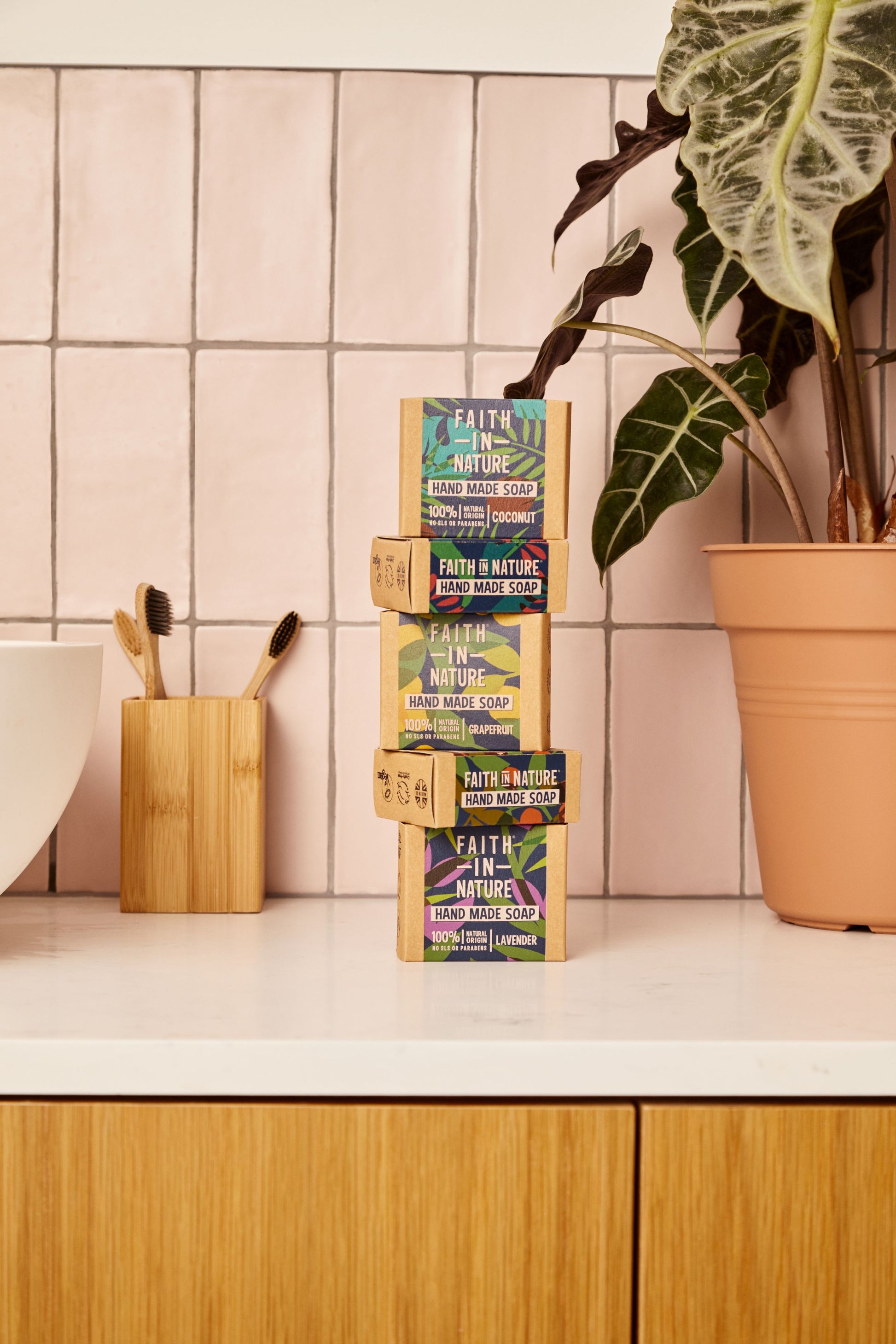 En stabel af fire Faith In Nature sæbebarer i deres 100% genanvendelige papemballage med varianterne Coconut, Grapefruit og Lavender på et badeværelse.