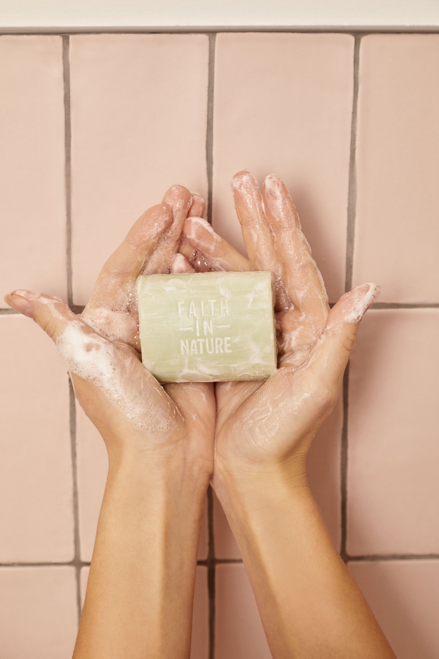 Hænder der vaskes med en lysegrøn Faith In Nature sæbebar, som danner et blødt og naturligt skum mod en baggrund af lyserøde fliser.